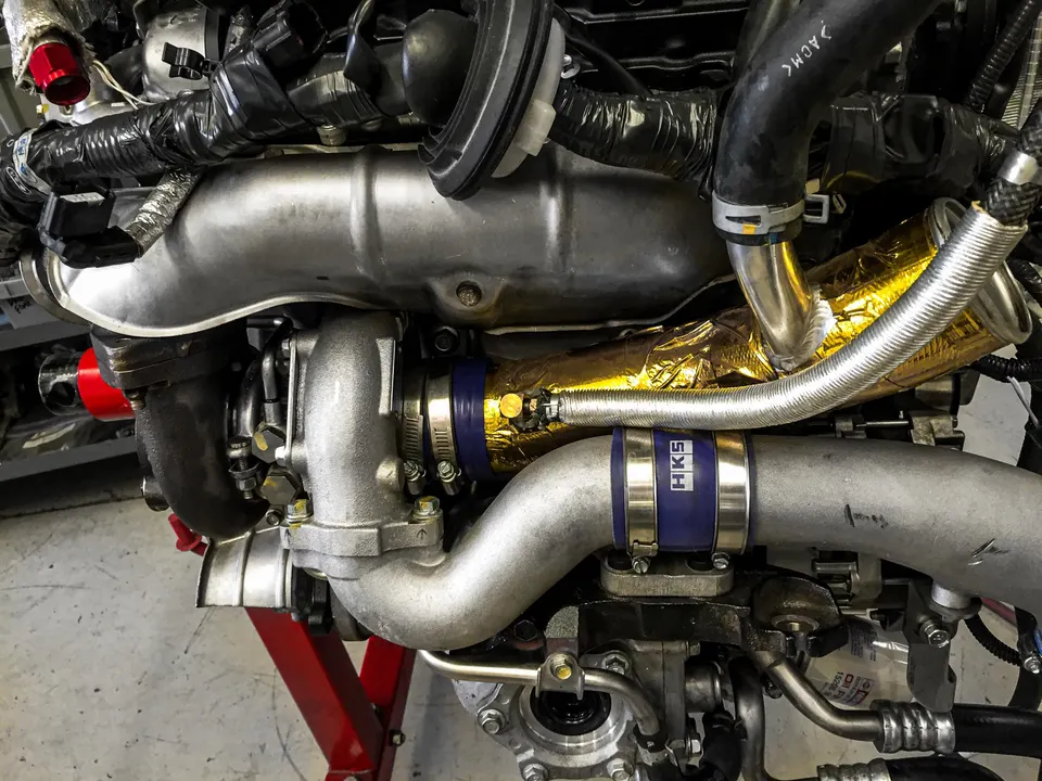 Close-up of a performance engine bay featuring a turbocharger, boost piping with HKS coupler, and gold heat shielding.
