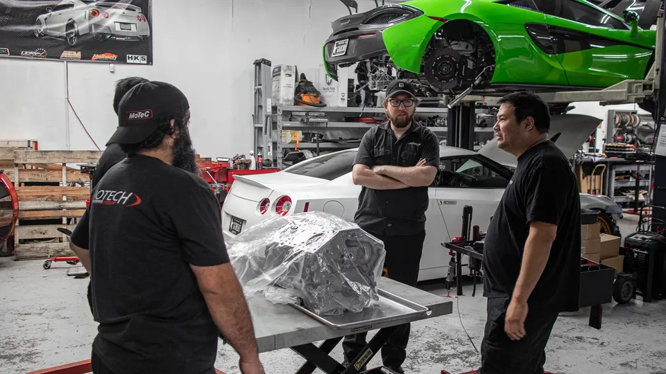 A group of mechanics discussing a large engine part in a performance tuning shop with a green McLaren and white Nissan GT-R.