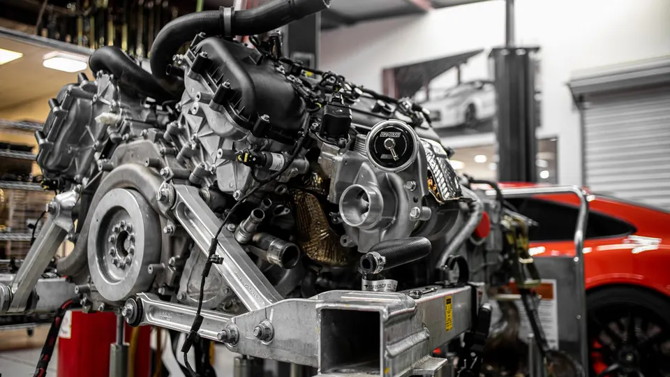 High-performance engine with a prominent turbocharger mounted on a stand in a workshop, with a red Porsche in the background.