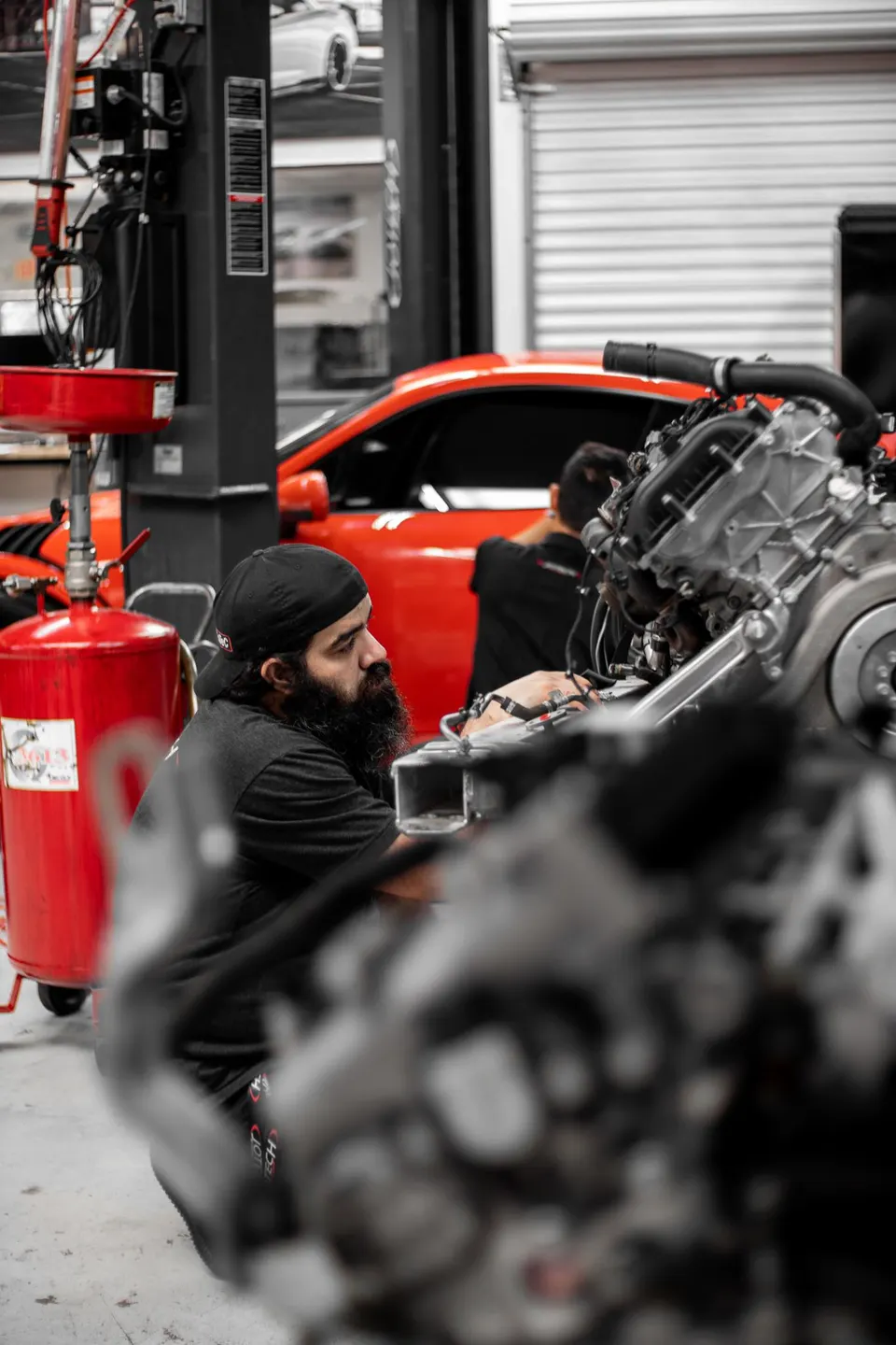 Mechanic working on a car engine in a performance tuning shop with a red car on a lift in the background.