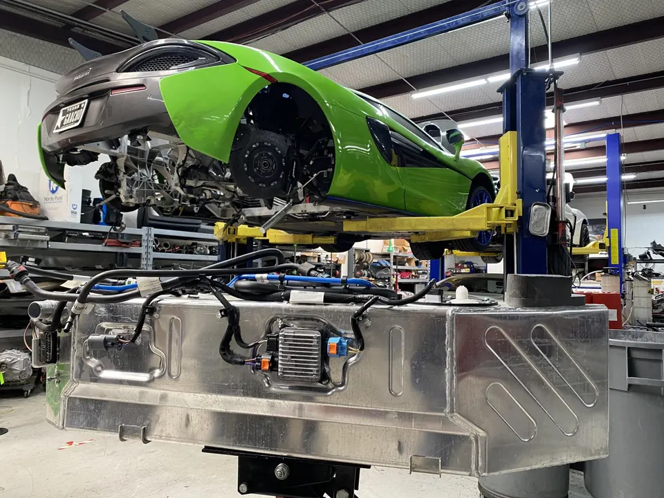 A green and grey McLaren supercar on a lift in a performance shop, with its fuel tank removed and visible in the foreground.