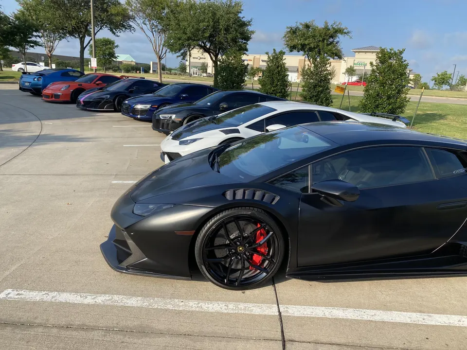 A line of supercars including a black Lamborghini Huracan, white Lamborghini, Audi R8, McLaren, Porsche, and Nissan GT-R parked at a car meet.
