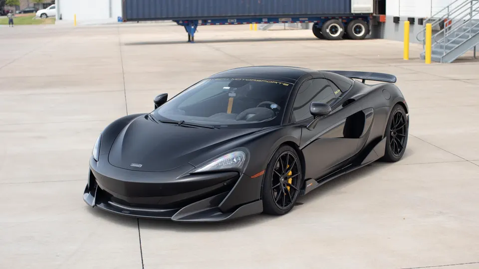 Black McLaren supercar with aftermarket wheels, carbon fiber front splitter, and rear wing parked on a concrete lot.