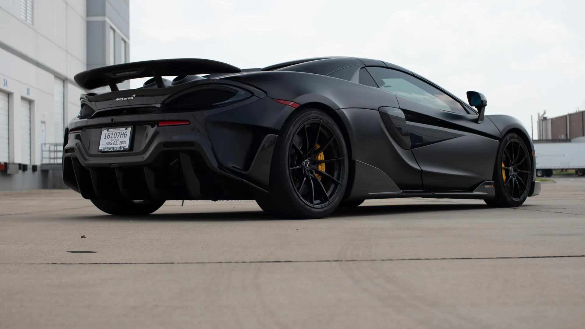 A black McLaren supercar with a large rear wing and aggressive diffuser parked in front of loading docks.