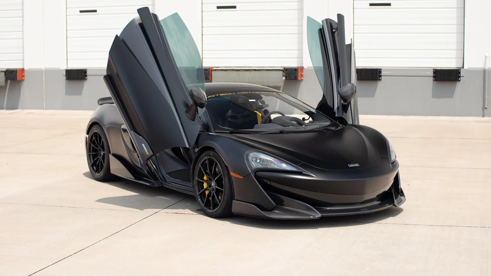 Matte black McLaren supercar with butterfly doors open, parked in front of white garage doors.