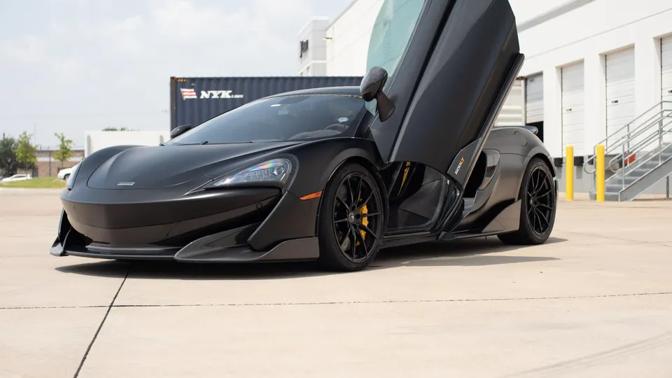 A black McLaren 600LT supercar with butterfly doors open, parked in an industrial area.