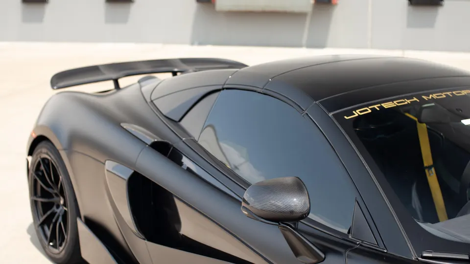 Matte black McLaren supercar with carbon fiber rear wing and side mirror, featuring Jotech Motorsports branding.