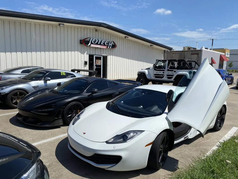 Multiple high-performance cars including a white McLaren, black McLaren, silver Nissan GT-R, and white Jeep parked outside Jotech Motorsports.