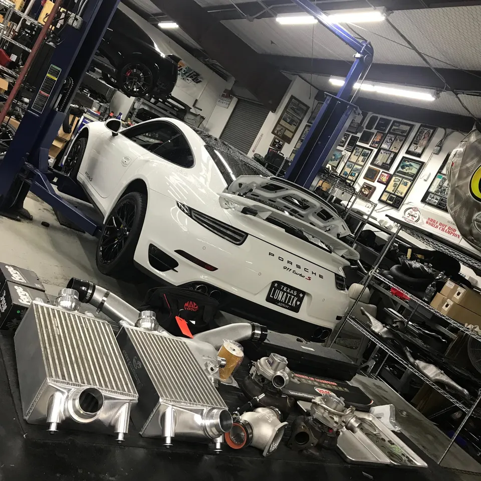 A white Porsche 911 Turbo S on a lift in a performance shop with turbochargers and intercoolers on a table.