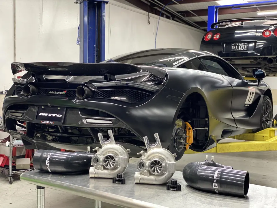 McLaren 720S on a lift in a performance shop with new turbochargers and boost piping on a table, a Nissan GT-R is also on a lift in the background.