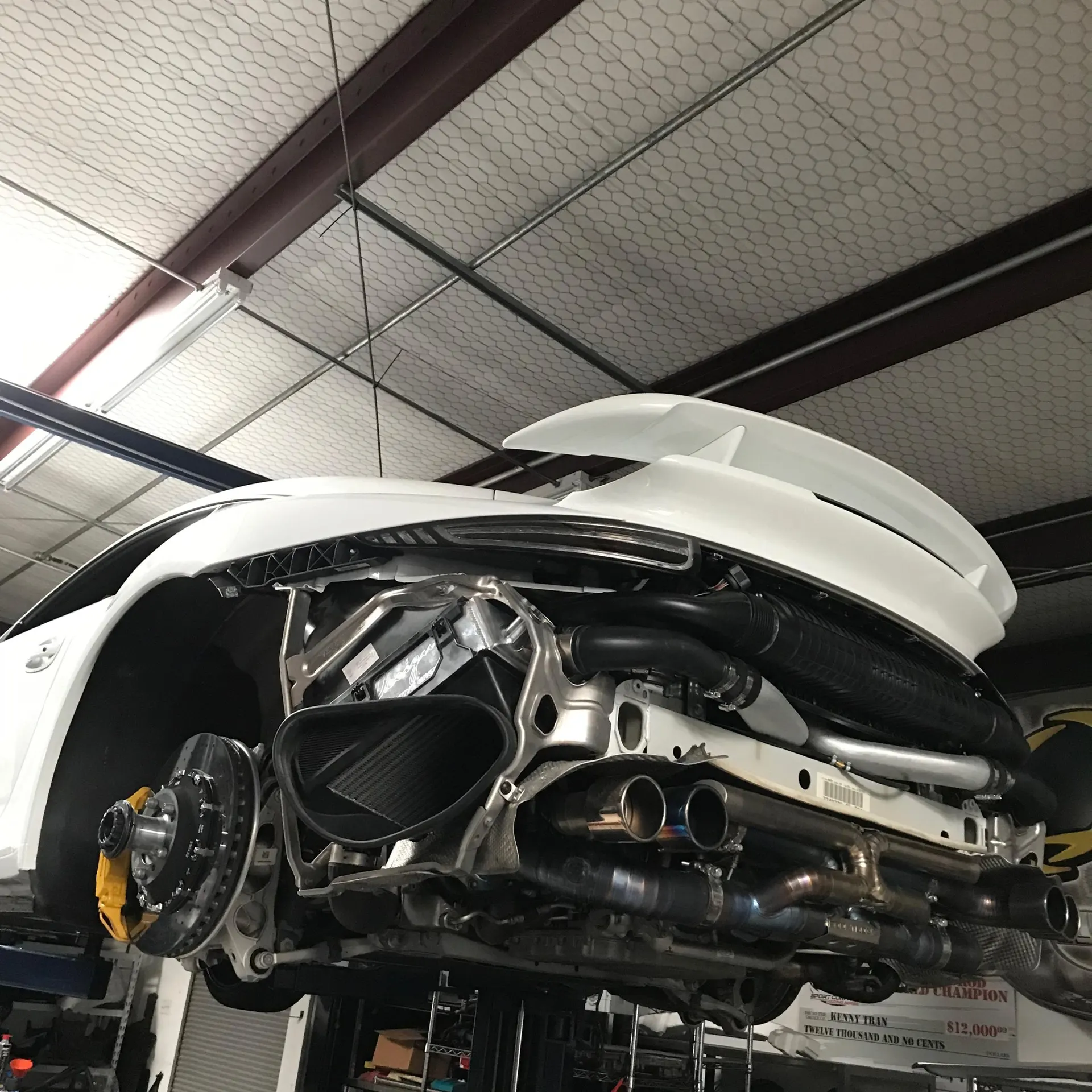 A white Porsche 911 GT3 RS on a lift in a performance shop, with its rear bumper removed to reveal the aftermarket exhaust and turbo system.