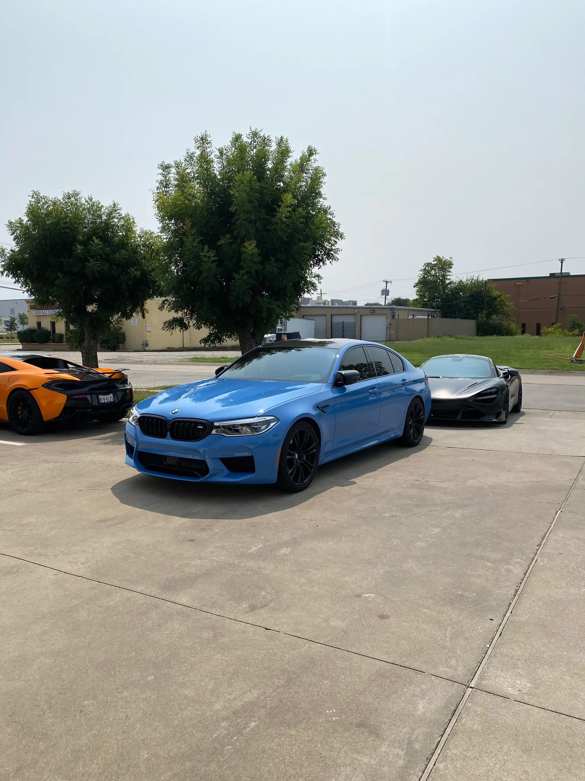 Blue BMW M5 sedan parked between an orange McLaren and a grey McLaren supercar in an outdoor lot.