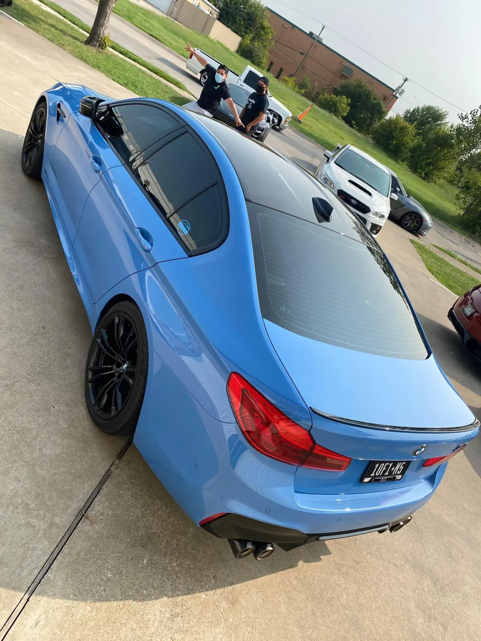 Blue BMW M5 with black wheels and aftermarket exhaust parked on a driveway with people and other cars in the background.