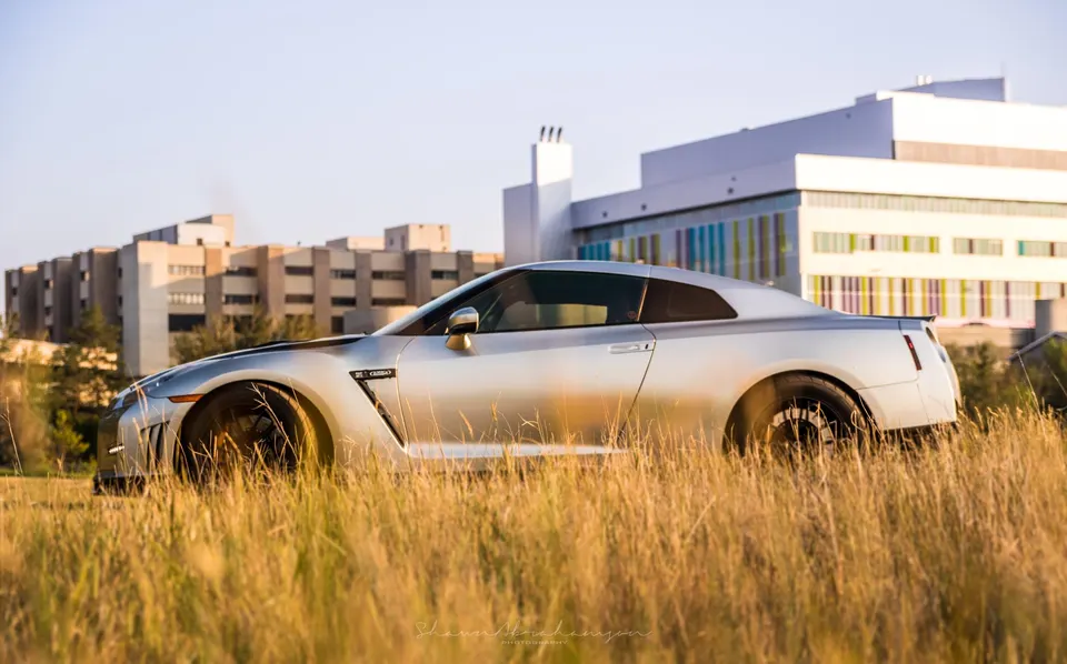 Silver Nissan GT-R parked in a field of tall golden grass with modern buildings in the background under a clear sky.