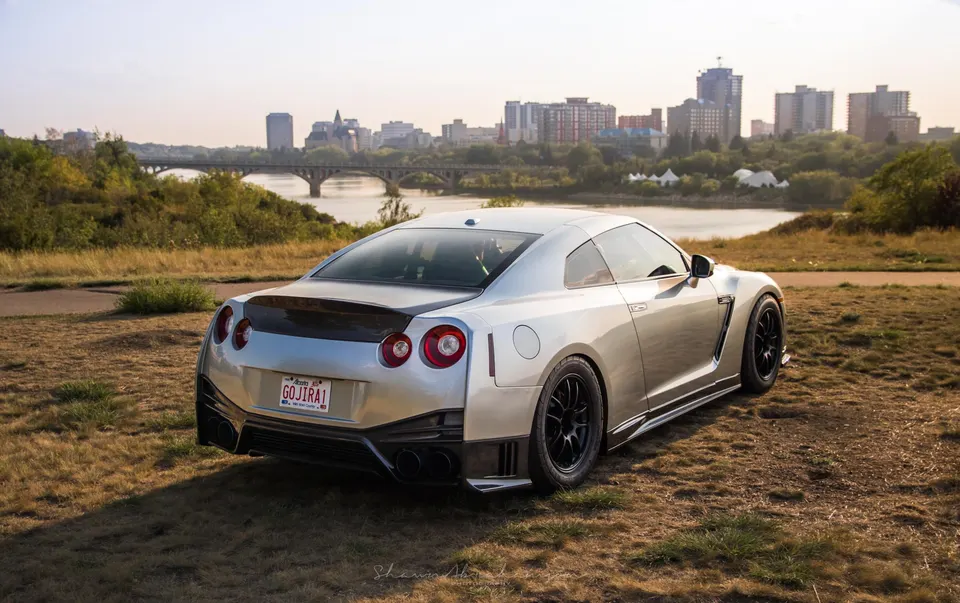Silver Nissan GT-R R35 with aftermarket wheels parked on a grassy hill overlooking a city skyline and river.