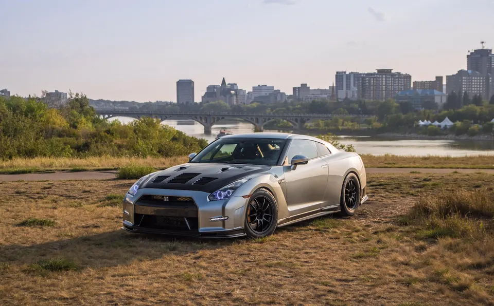 A silver Nissan GT-R with a carbon fiber hood and aftermarket wheels parked in a grassy field with a city skyline in the background.