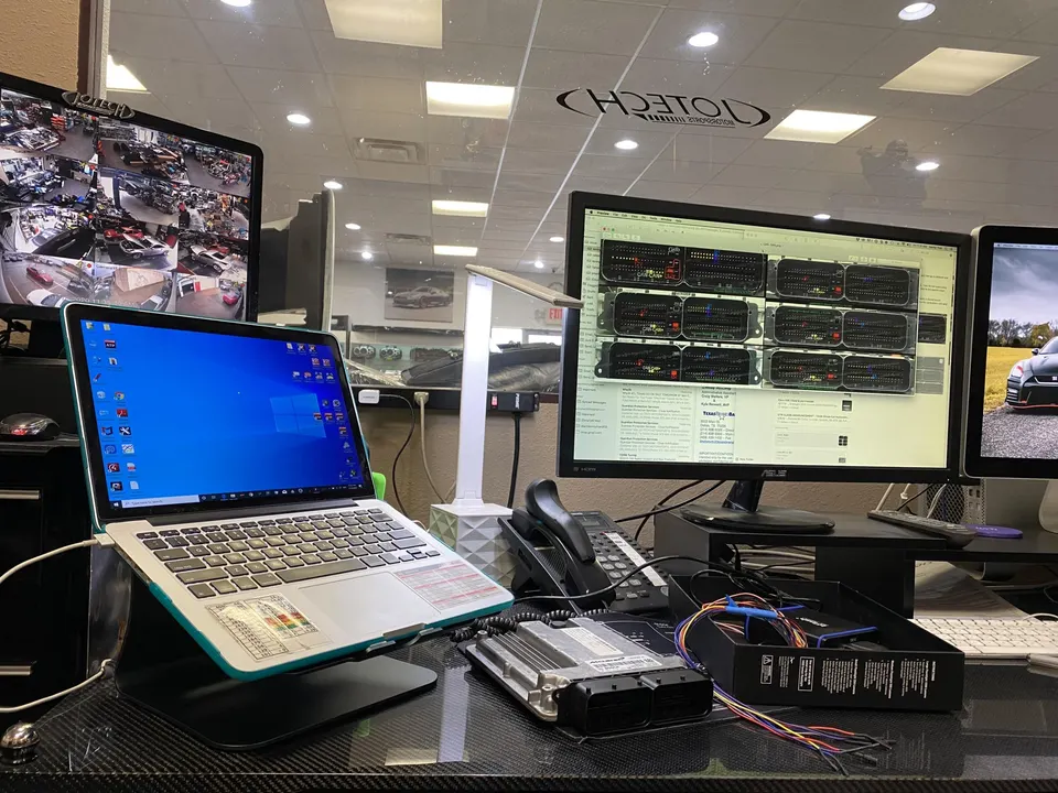A shop desk with a laptop, multiple monitors displaying tuning software and security feeds, and an ECU with wiring.
