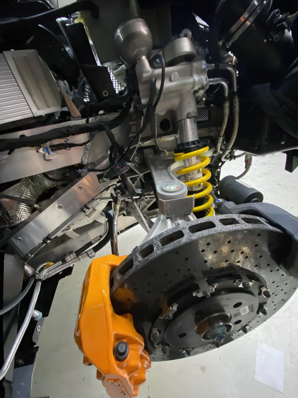 Close-up of a McLaren's front suspension and braking system, featuring a yellow coilover, large brake rotor, and orange caliper.