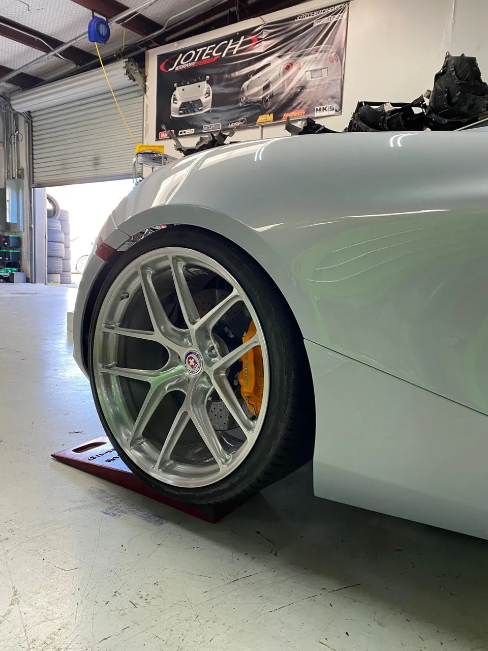 Close-up of a silver sports car's front wheel featuring multi-spoke silver HRE wheels and yellow brake calipers in a shop.