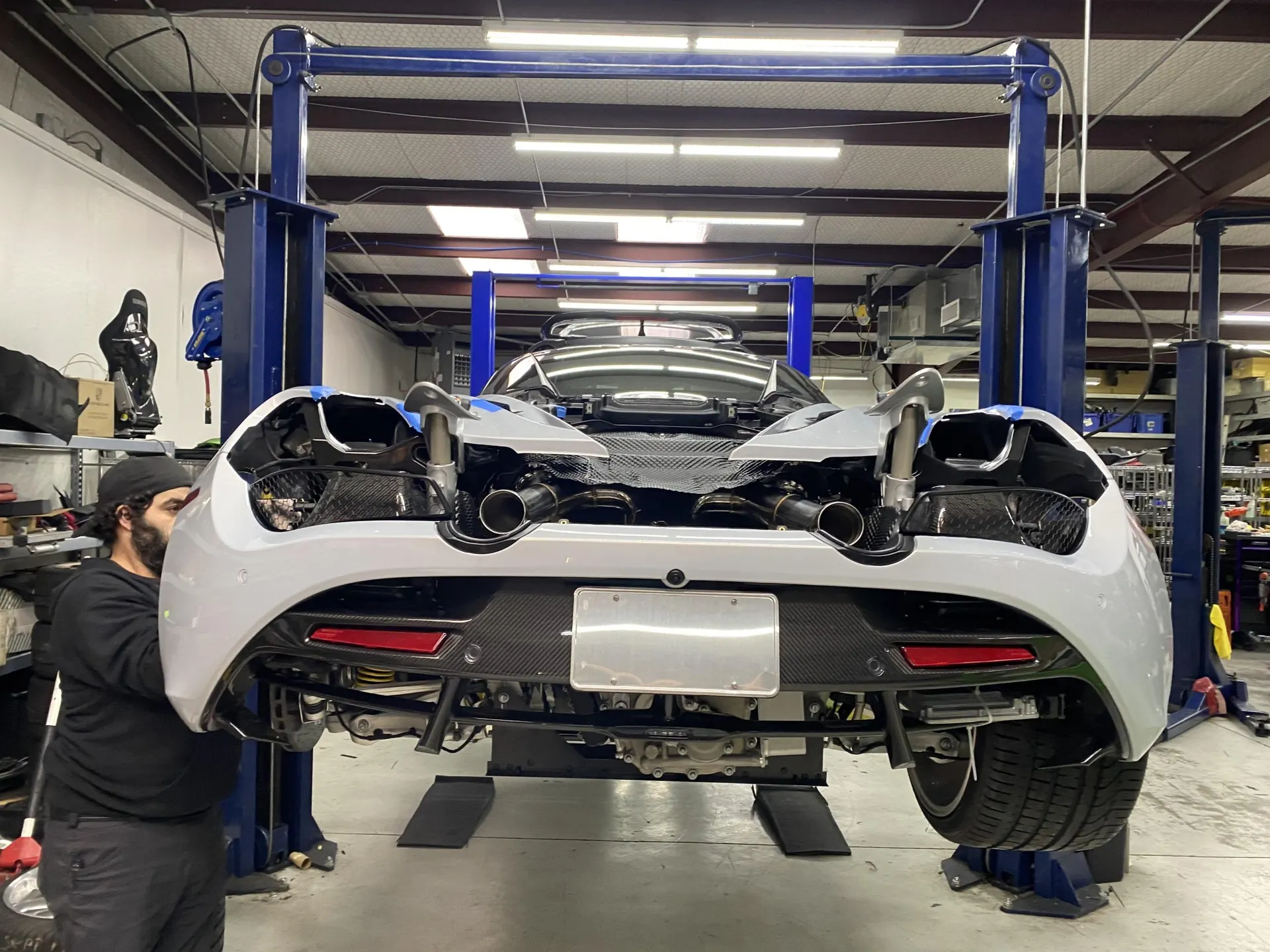 Rear view of a McLaren supercar on a lift in a shop with its rear bumper partially removed, revealing the exhaust system, as a mechanic works on it.