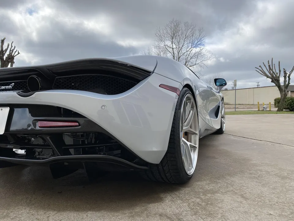 Rear-side view of a silver McLaren 720S with aftermarket multi-spoke wheels parked on concrete under a cloudy sky.