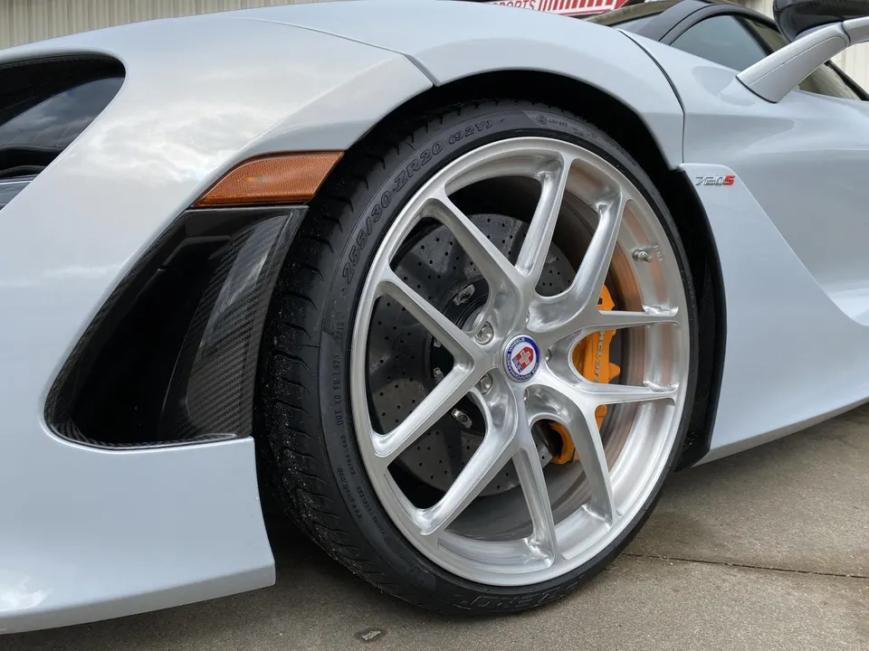 Close-up of a white McLaren 720S front wheel with silver HRE custom wheels, orange brake calipers, and carbon fiber fender trim.