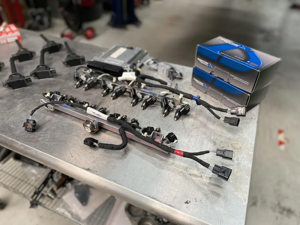 McLaren fuel injectors, fuel rails, ignition coils, and ECU laid out on a metal workbench in a performance tuning shop.