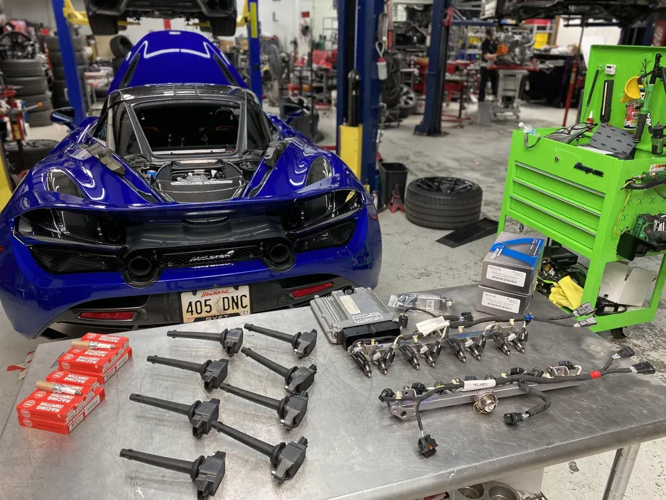 Blue McLaren 720S in a performance shop with fuel injectors, ECU, spark plugs, and ignition coils laid out on a workbench.