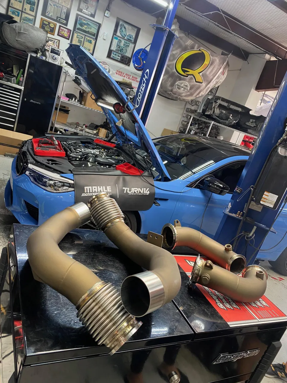 Blue BMW M2 on a lift in a Jotech Motorsports shop with performance exhaust downpipes on a toolbox in the foreground.