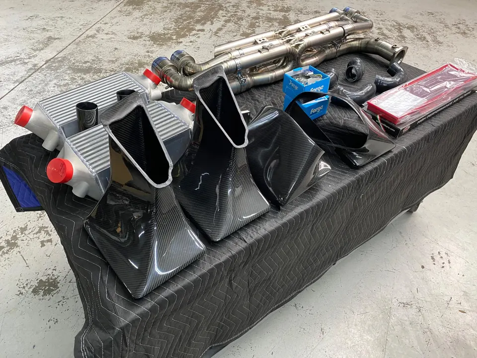 A collection of high-performance carbon fiber intake, intercoolers, and titanium exhaust parts laid out in a shop.