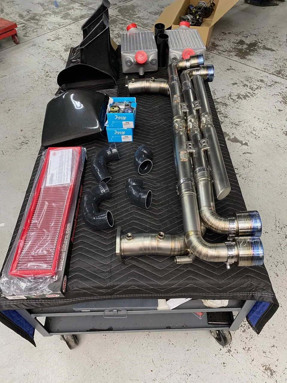 Aftermarket exhaust system, carbon fiber intake, and intercoolers laid out on a cart in a tuning shop.