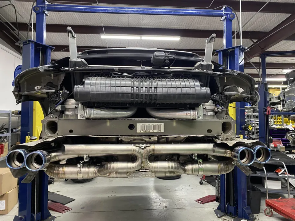 A Porsche 911 on a lift in a shop, showcasing an aftermarket titanium exhaust system with blue tips.