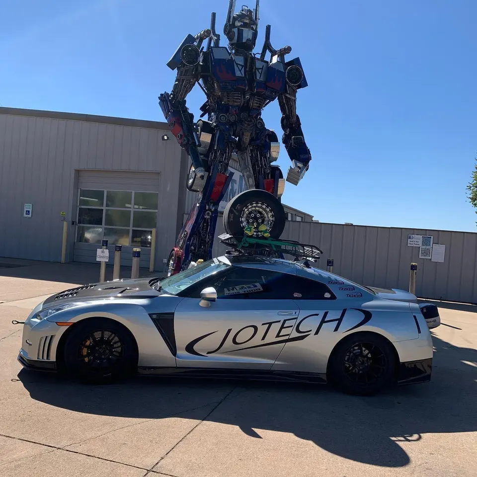 Silver Nissan GT-R with Jotech livery parked in front of a giant Optimus Prime statue, carrying a spare wheel on its roof rack.