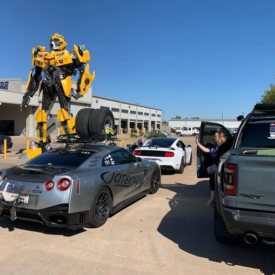 Nissan GT-R with drag tires on a roof rack and a Ford Mustang parked outside Jotech Motorsports with a Bumblebee statue.