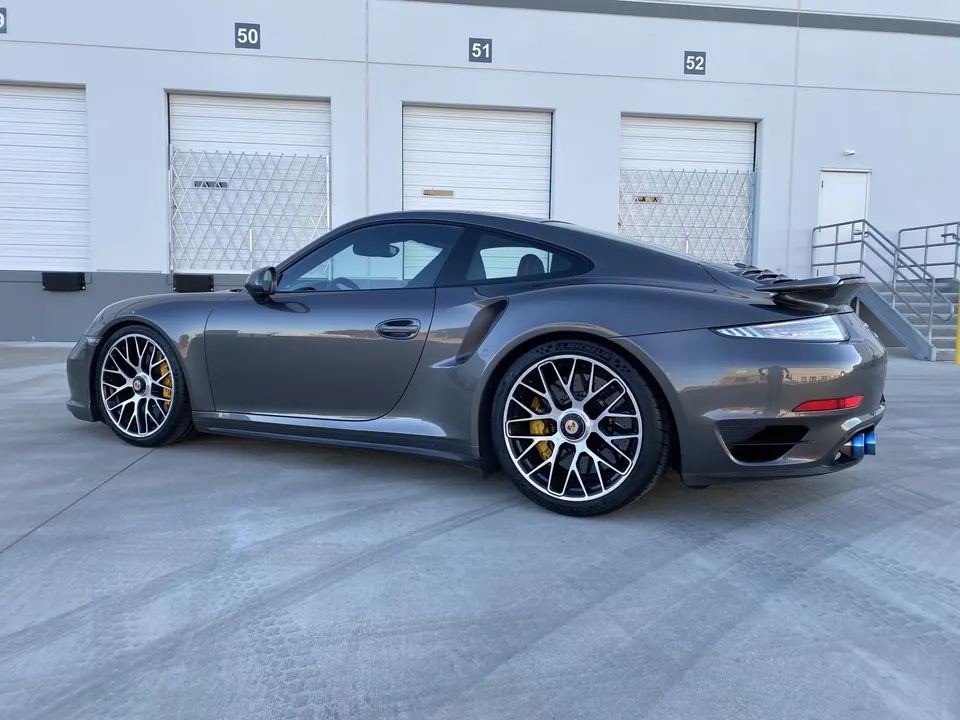 A grey Porsche 911 with aftermarket blue exhaust tips and custom wheels parked in front of a warehouse.