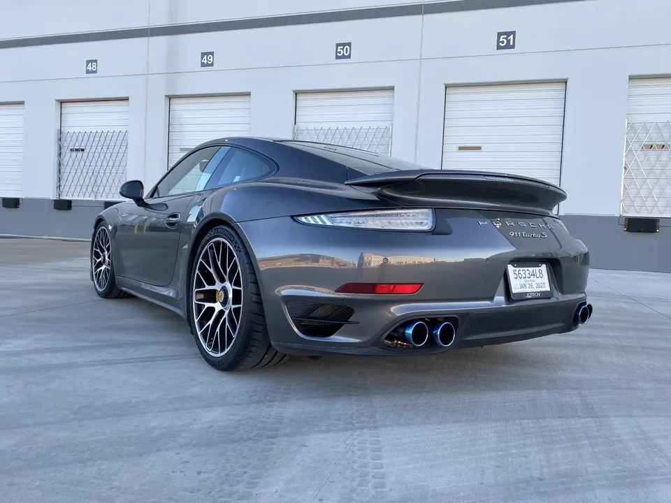 Rear view of a grey Porsche 911 Turbo S with aftermarket blue-tipped exhaust parked in front of a warehouse.