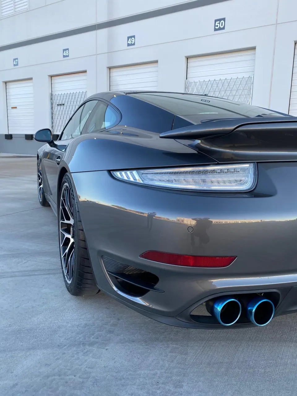 A dark grey Porsche 911 with aftermarket blue exhaust tips parked in front of numbered garage doors.