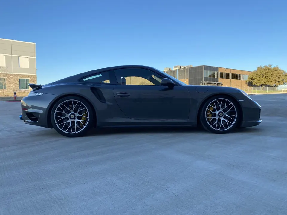 A grey Porsche 911 Turbo S parked outdoors with yellow brake calipers, showcasing its side profile.