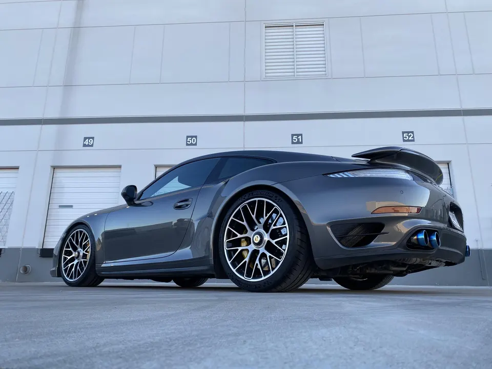 A grey Porsche 911 Turbo S with aftermarket wheels and blue exhaust tips parked in front of a building.