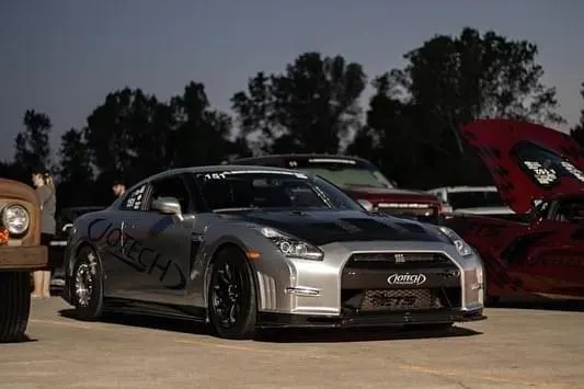A silver Nissan GT-R with a black hood and Jotech branding parked at a car event at night, with other vehicles and people in the background.