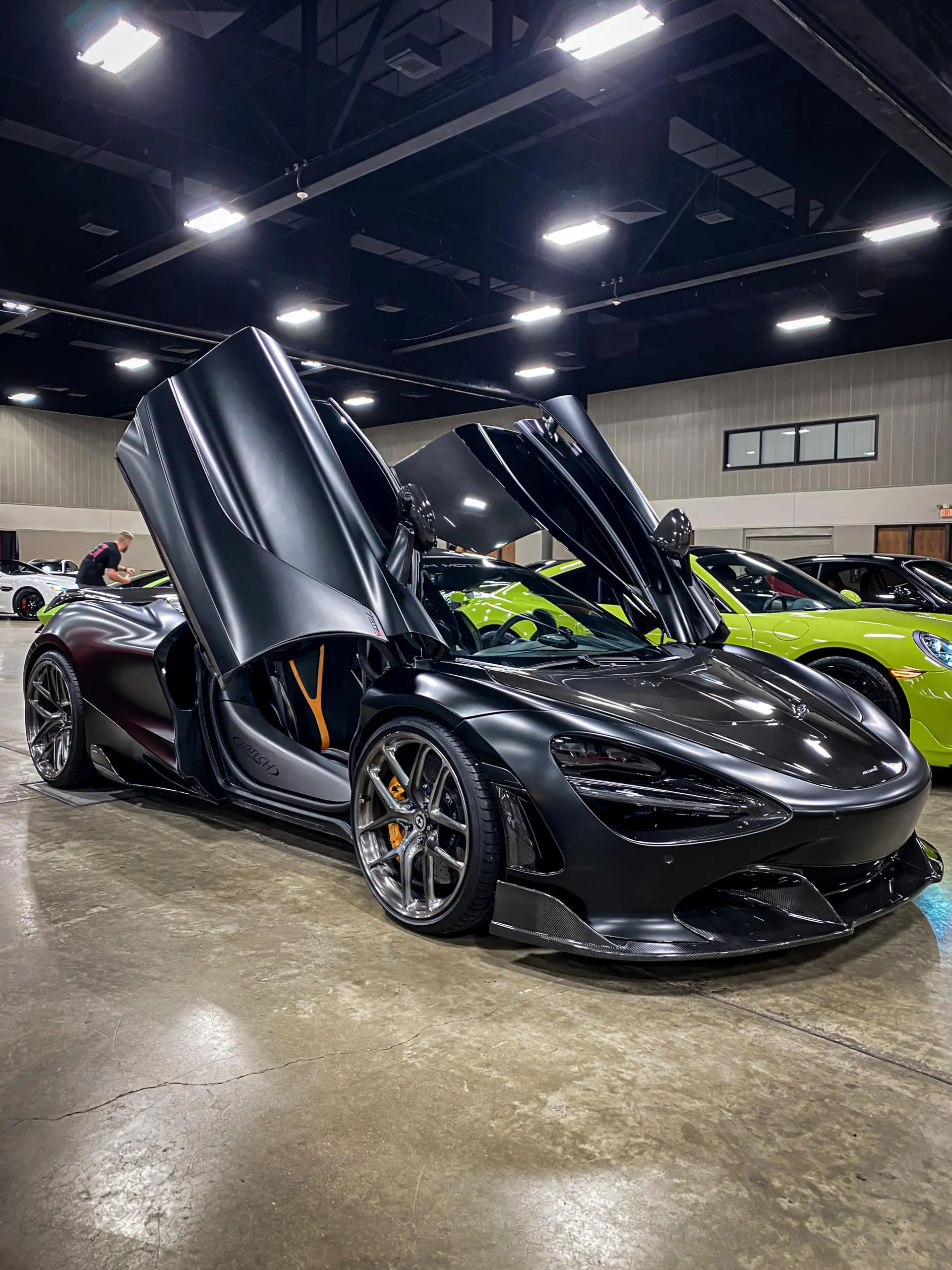 Matte black McLaren 720S with butterfly doors open, showcasing aftermarket wheels and carbon fiber aero at an indoor event.