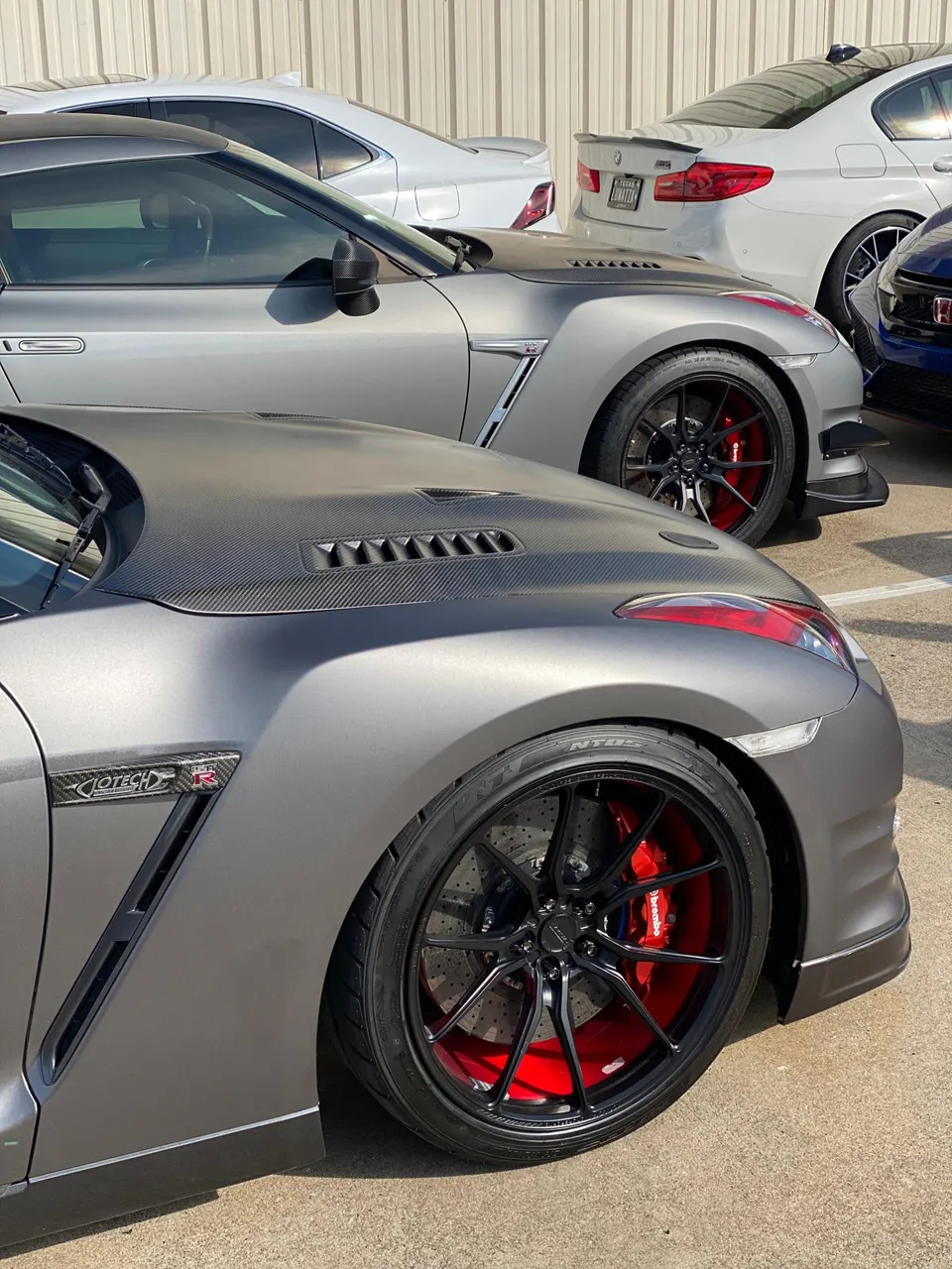 Two grey Nissan GT-Rs parked, showcasing custom black wheels with red Brembo calipers and carbon fiber hoods.