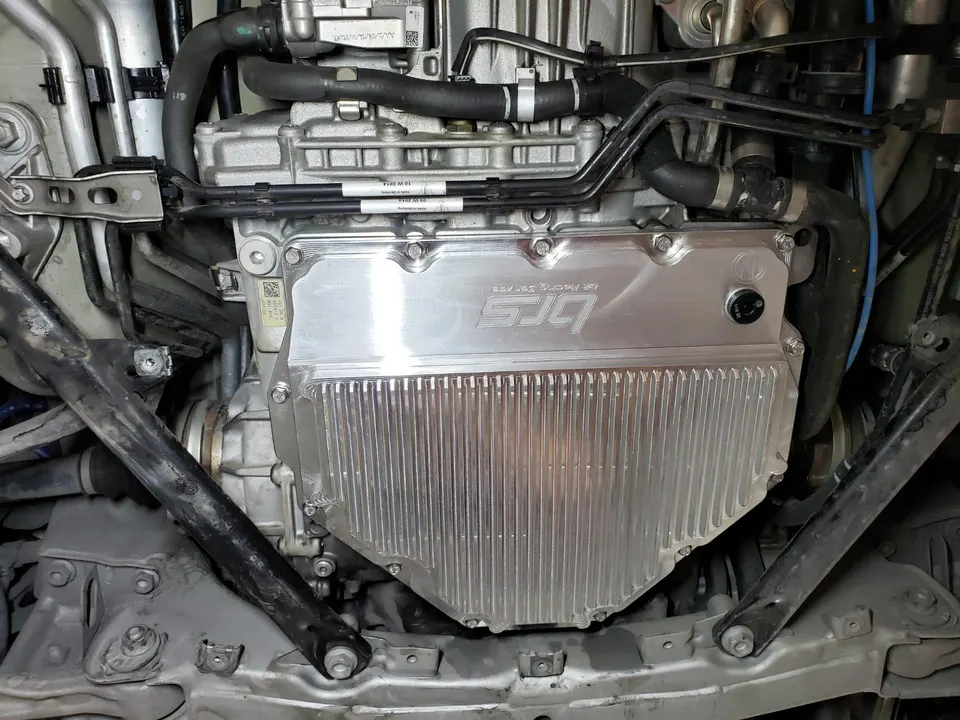 Close-up of an aftermarket SJQ performance oil pan installed on a car's engine in a shop.