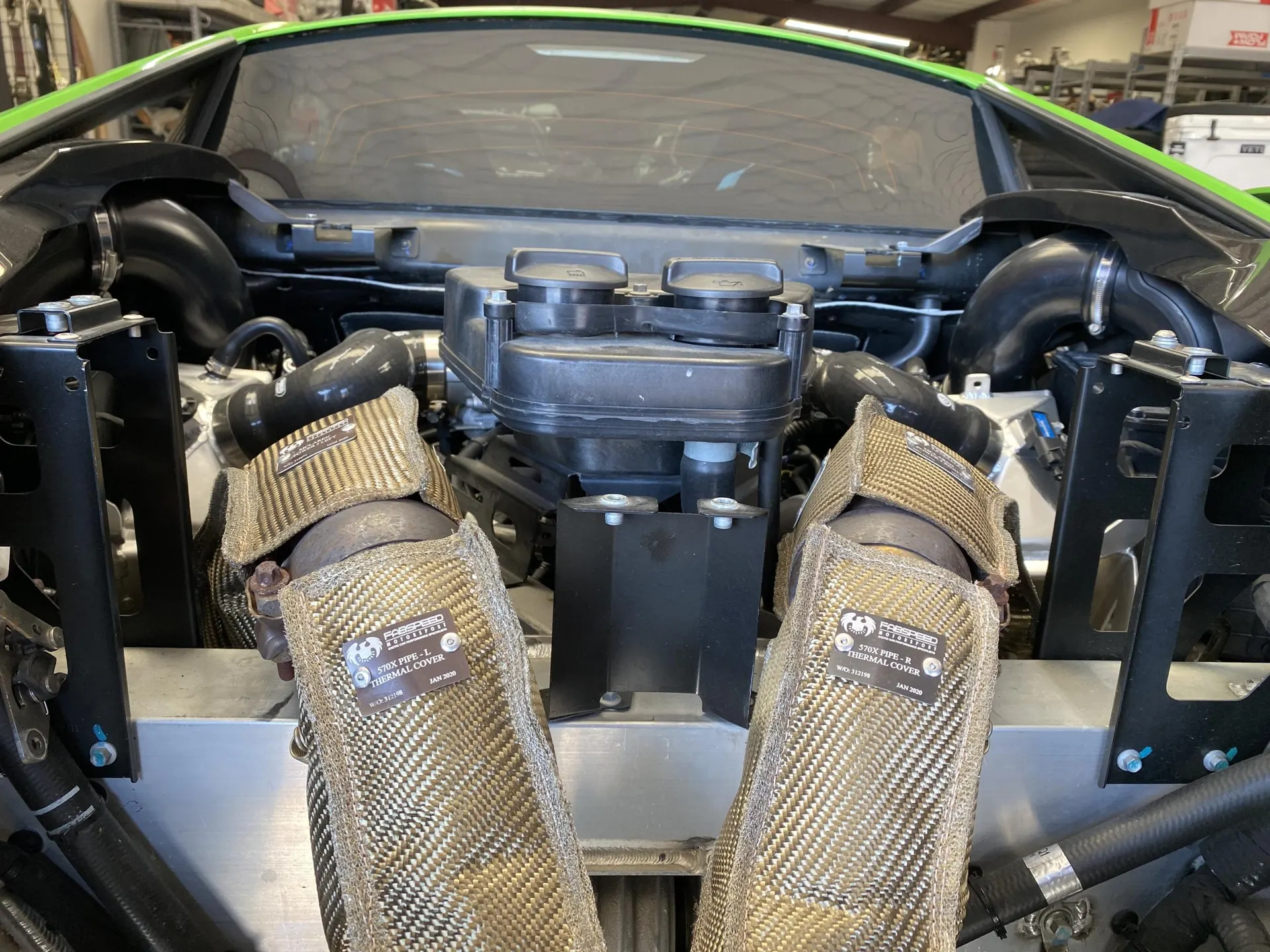 Close-up view of a Lamborghini engine bay showing aftermarket exhaust downpipes with thermal covers installed.