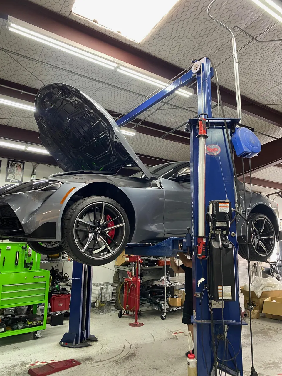 A gray Toyota Supra MK5 on a hydraulic lift in a performance tuning shop with its hood open, being worked on by a mechanic.