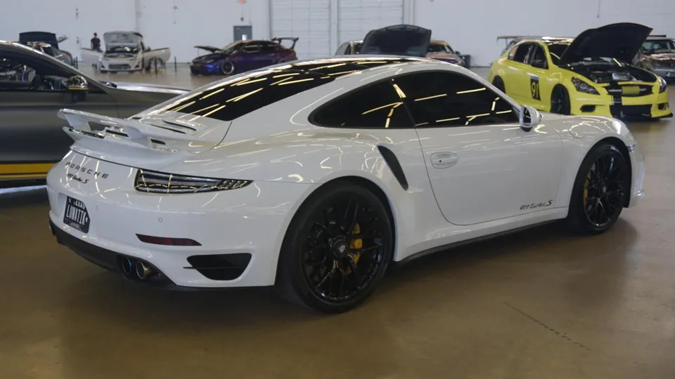 White Porsche 911 Turbo S with a rear wing and black wheels parked at an indoor car show with other modified vehicles.