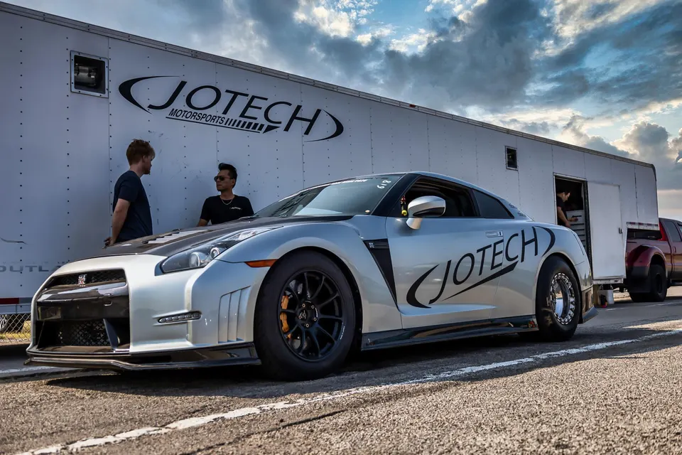 A silver Nissan GT-R drag car with Jotech Motorsports branding parked next to a Jotech trailer at a track event.