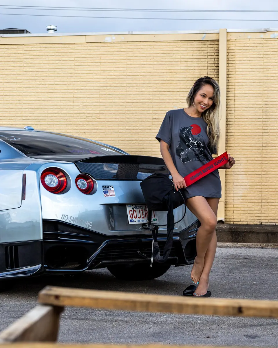 A woman in a grey t-shirt stands next to a silver Nissan GT-R with a carbon fiber trunk and HKS sticker.