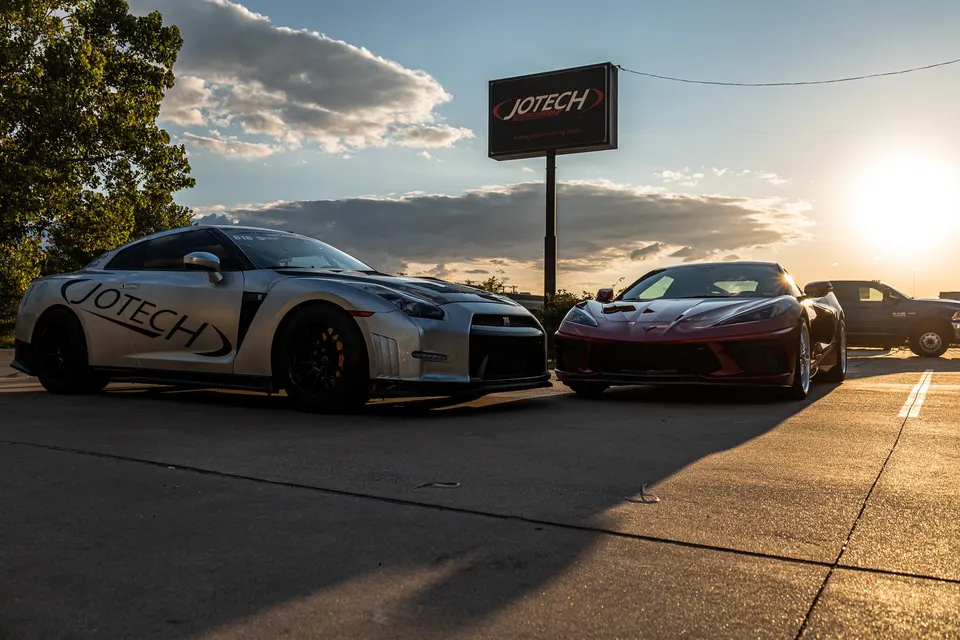 A silver Nissan GT-R and a red C8 Corvette parked side-by-side in front of the Jotech Motorsports sign at sunset.