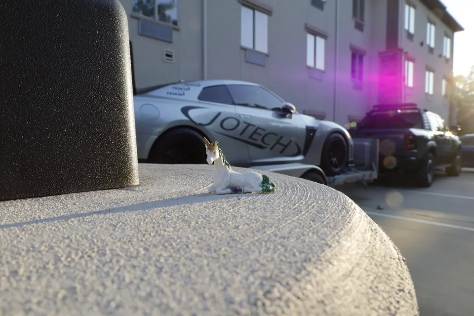 A silver Jotech Nissan GT-R on a car hauler, towed by a pickup truck, with a small unicorn figurine in the foreground.